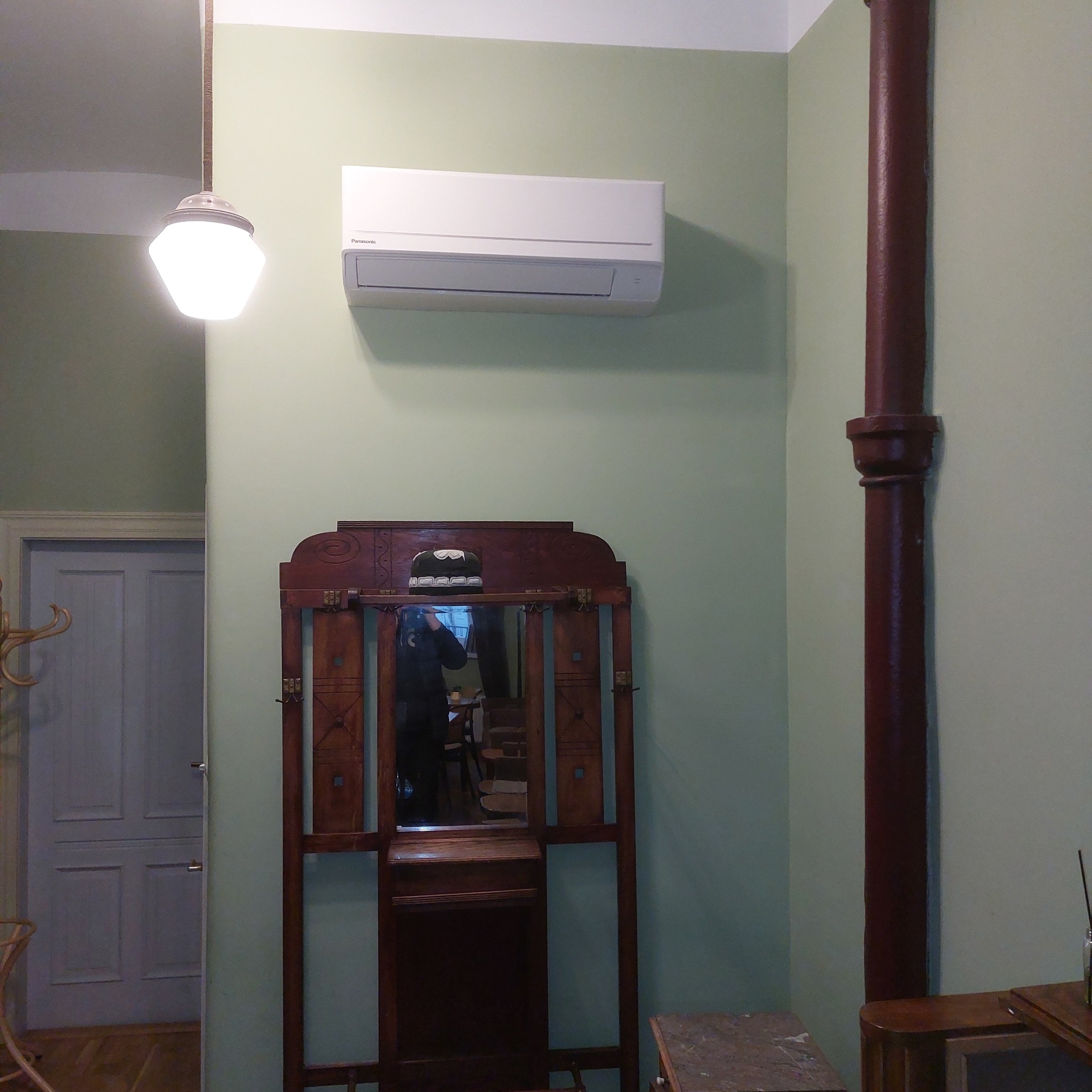 Biała klimatyzacja Panasonic zamontowana na zielonej ścianie w pomieszczeniu z elementami w stylu retro: drewniany wieszak z lustrem i brązowa rura pionowa.