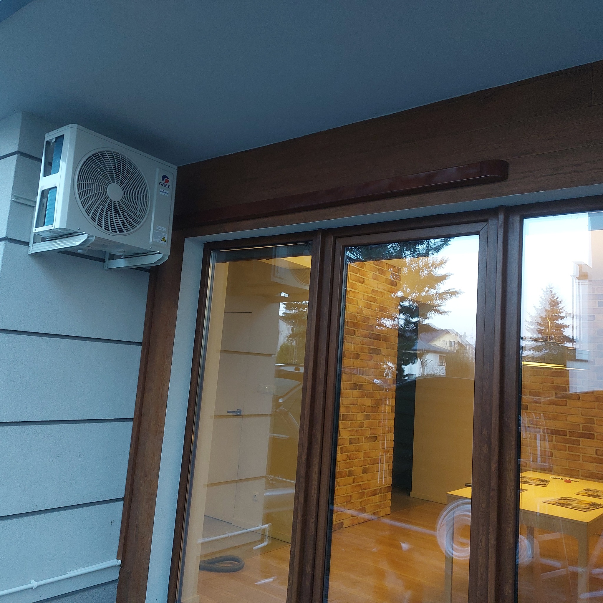 Zewnętrzna jednostka klimatyzacji marki Gree zamontowana na elewacji budynku obok okna z brązową ramą, widoczne odbicie drzewa w szybie.