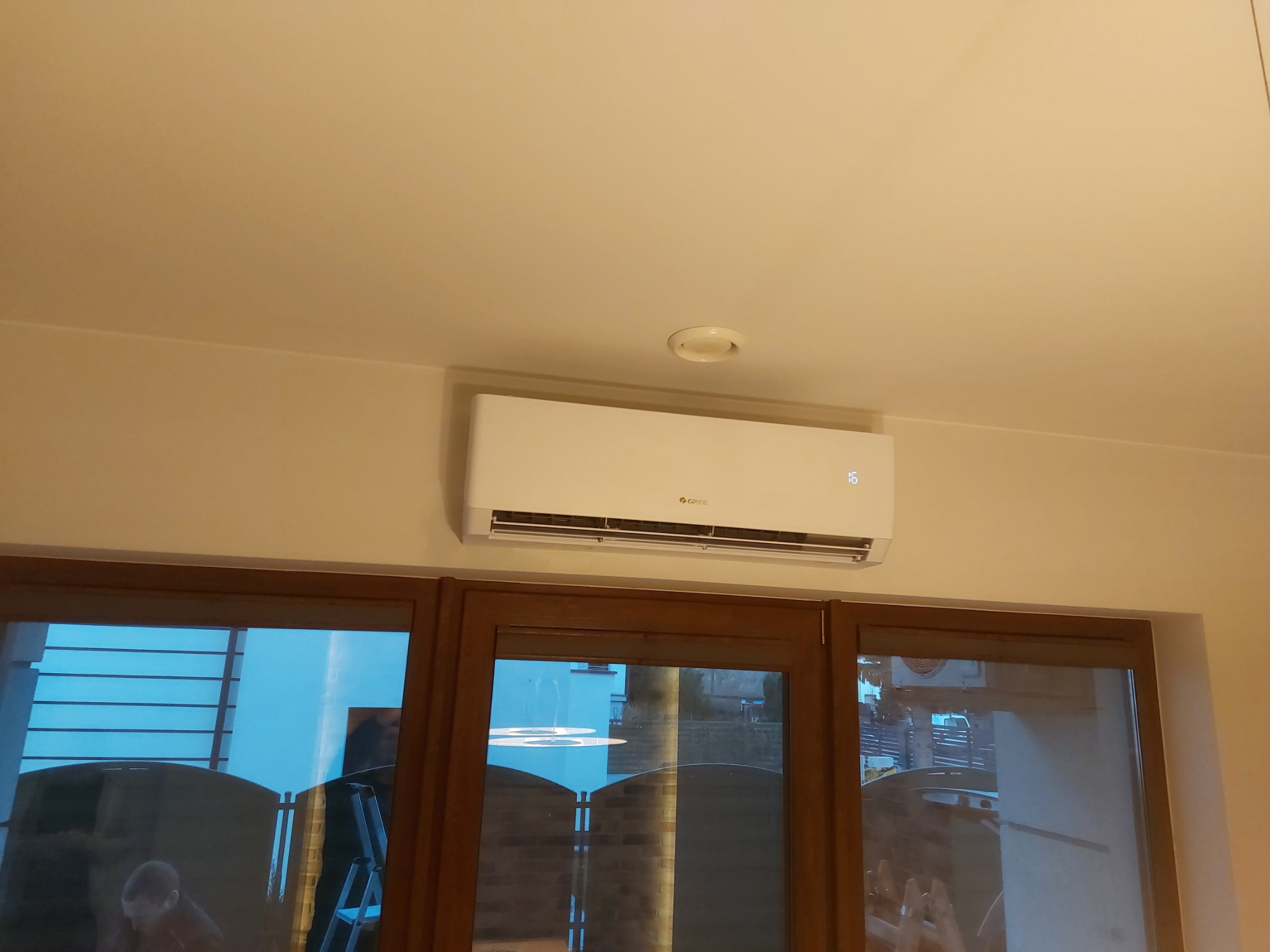 Biały klimatyzator marki Gree zamontowany na ścianie nad oknem z ciemnymi ramami, widoczny wyświetlacz temperatury i logo producenta.