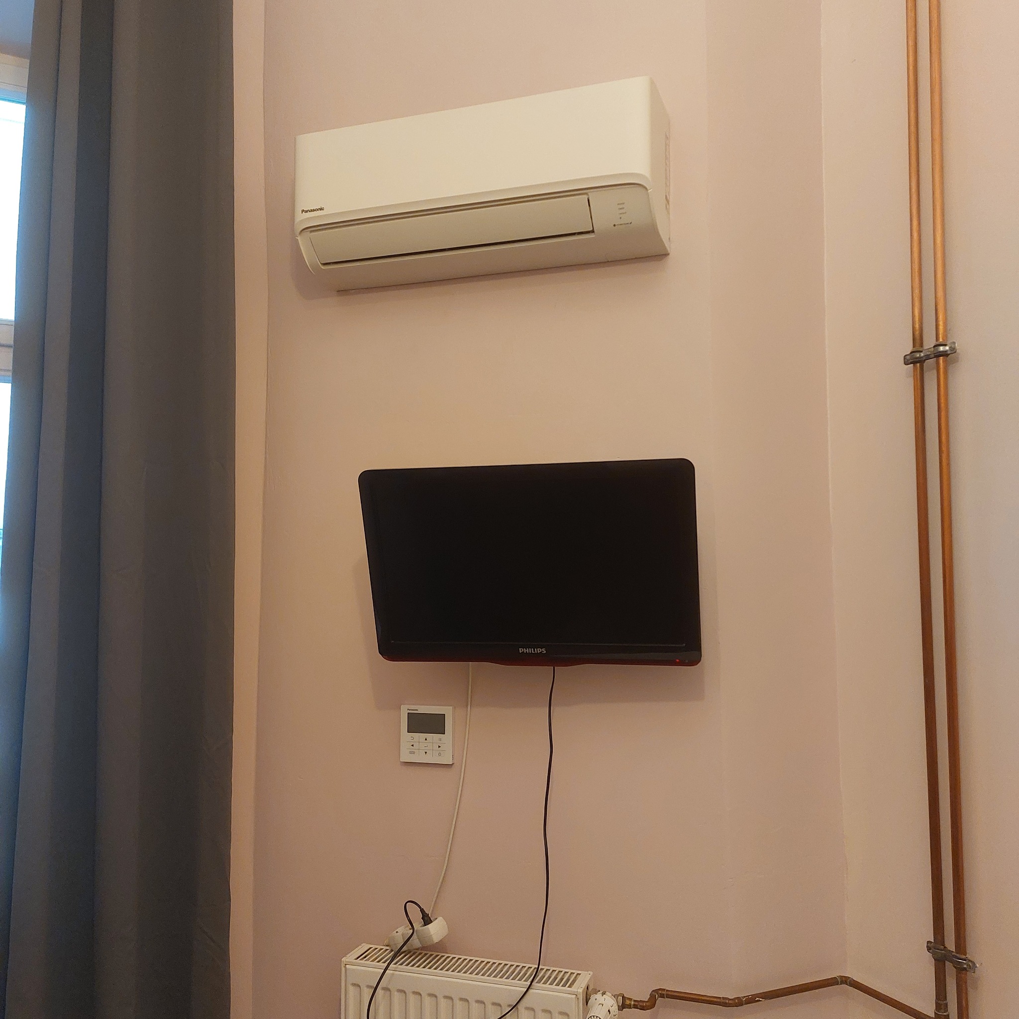 Biały klimatyzator Panasonic zamontowany na ścianie nad telewizorem Philips, z widocznym sterownikiem na ścianie, grzejnikiem i miedzianymi rurami instalacji grzewczej.