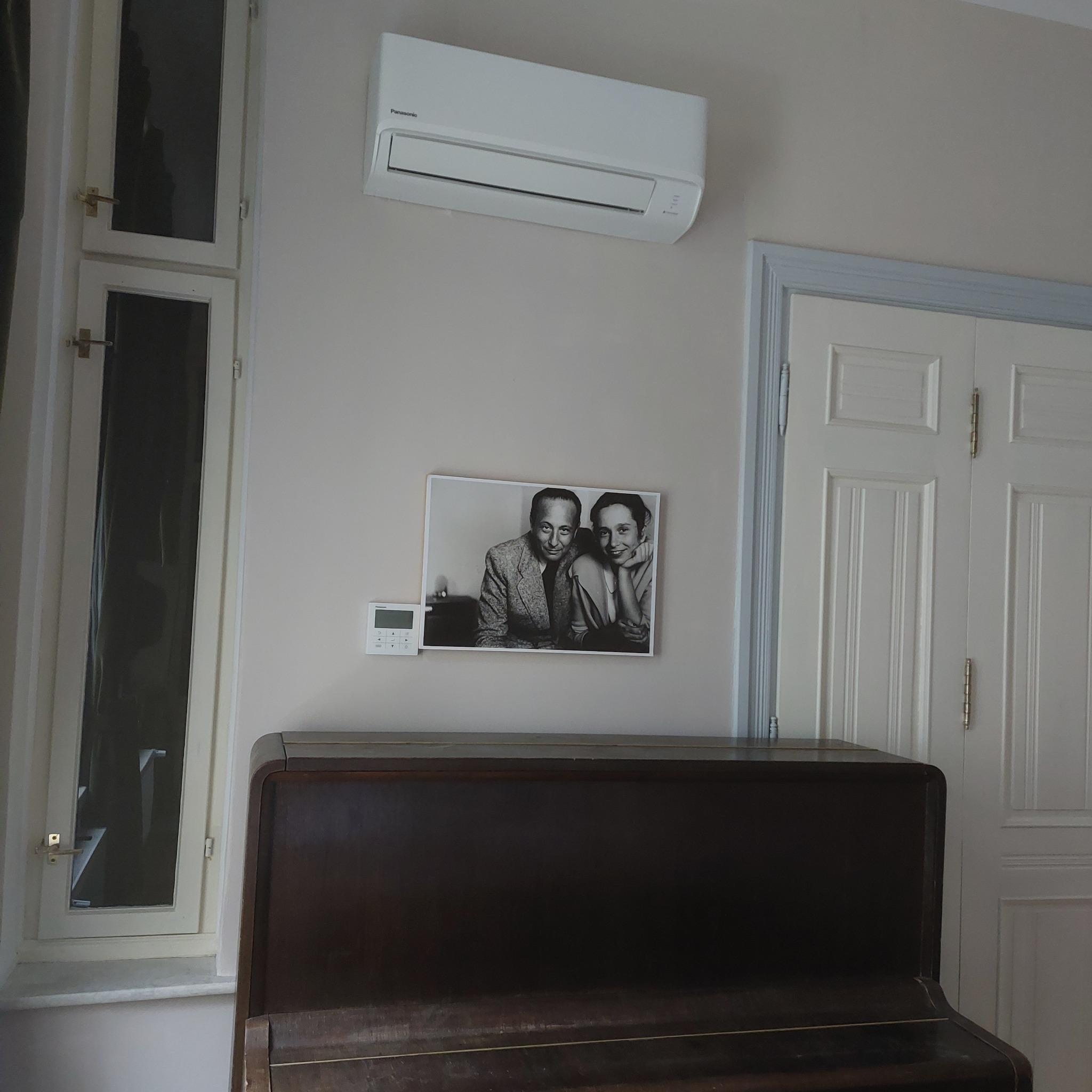 Biały klimatyzator Panasonic zamontowany nad czarno-białym zdjęciem pary i panelem sterowania, wiszący nad ciemnym pianinem w pokoju z oknem i białymi drzwiami.