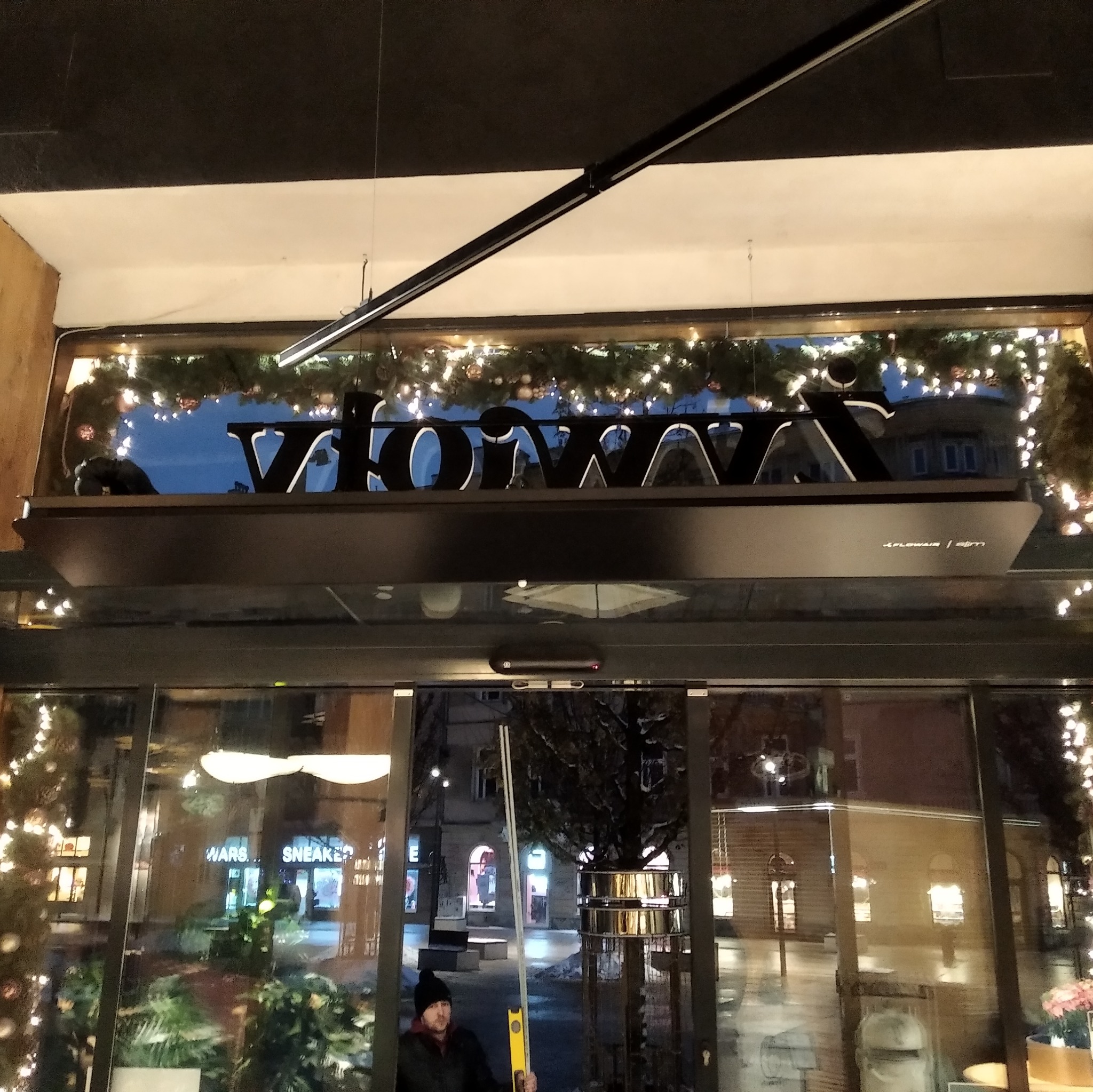 Instalator z poziomicą ustawia logo 'XFLOWAIR / slim' klimatyzacji nad wejściem do budynku udekorowanego świątecznymi lampkami.