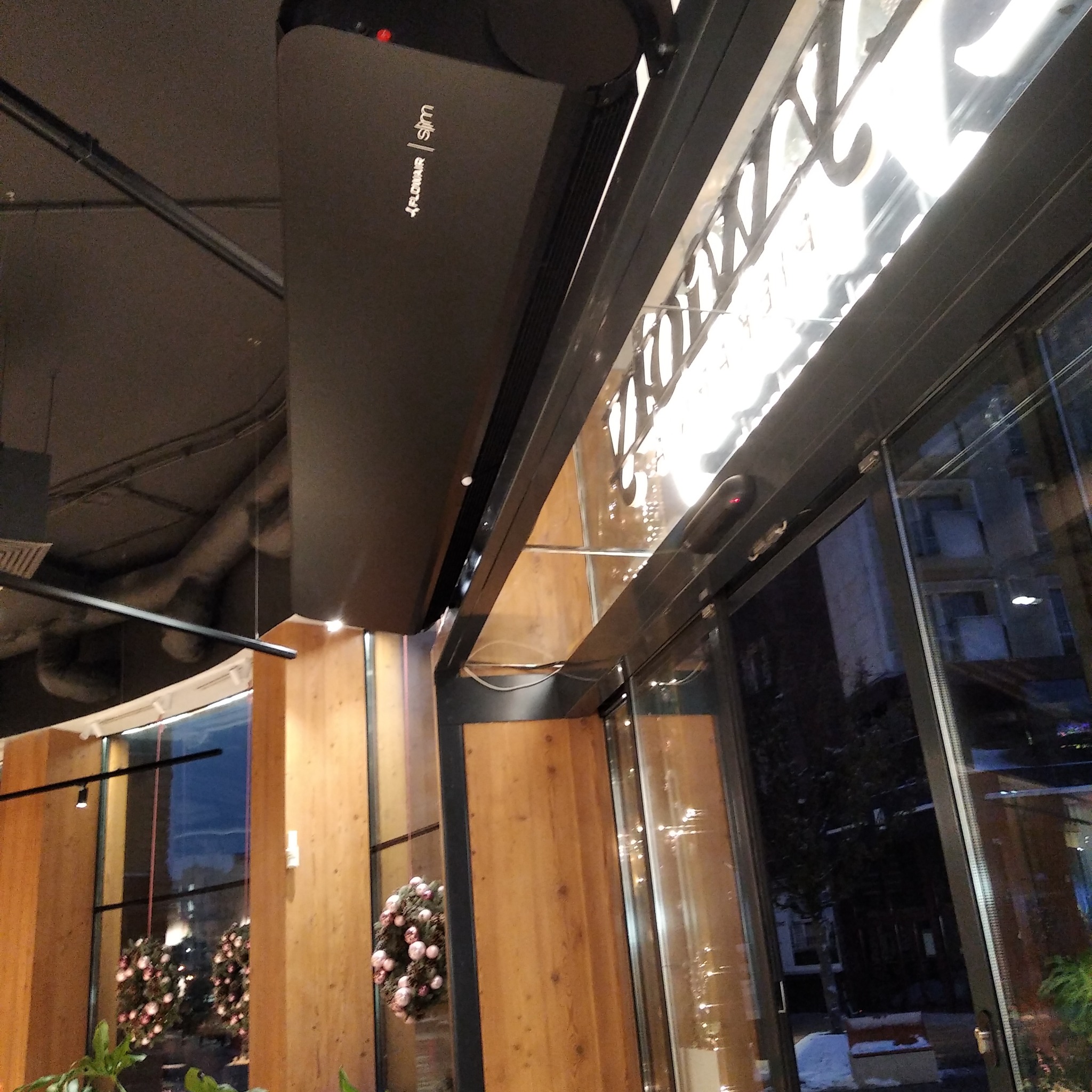 Czarna jednostka klimatyzacyjna Flowair Slim zamontowana pod sufitem w lokalu komercyjnym, widoczne elementy instalacji i oświetlenie sufitowe.