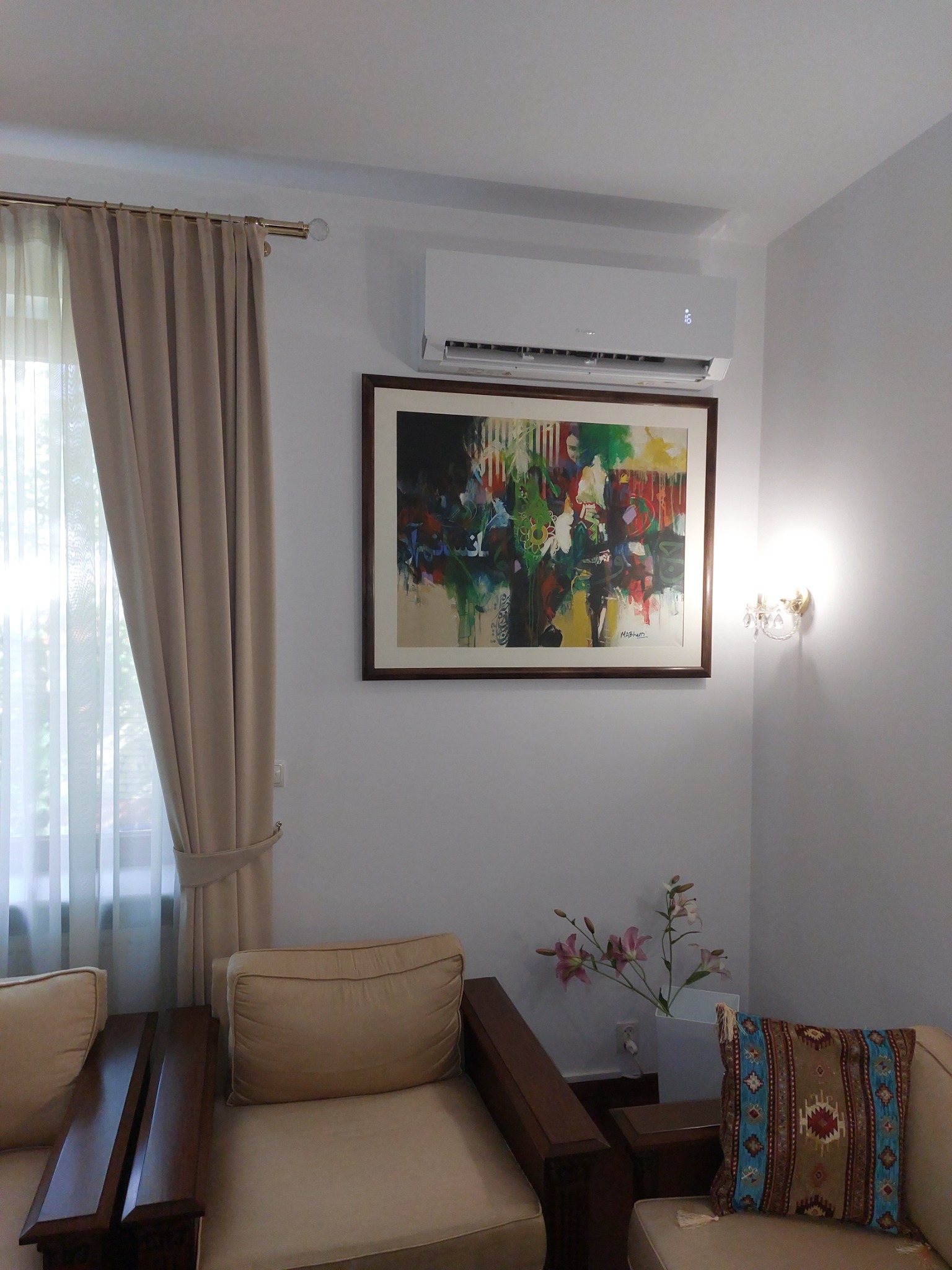 Nowoczesny klimatyzator ścienny w jasnym salonie, zawieszony nad abstrakcyjnym obrazem w drewnianej ramie, widoczne beżowe zasłony, drewniane meble i kwiaty w wazonie.