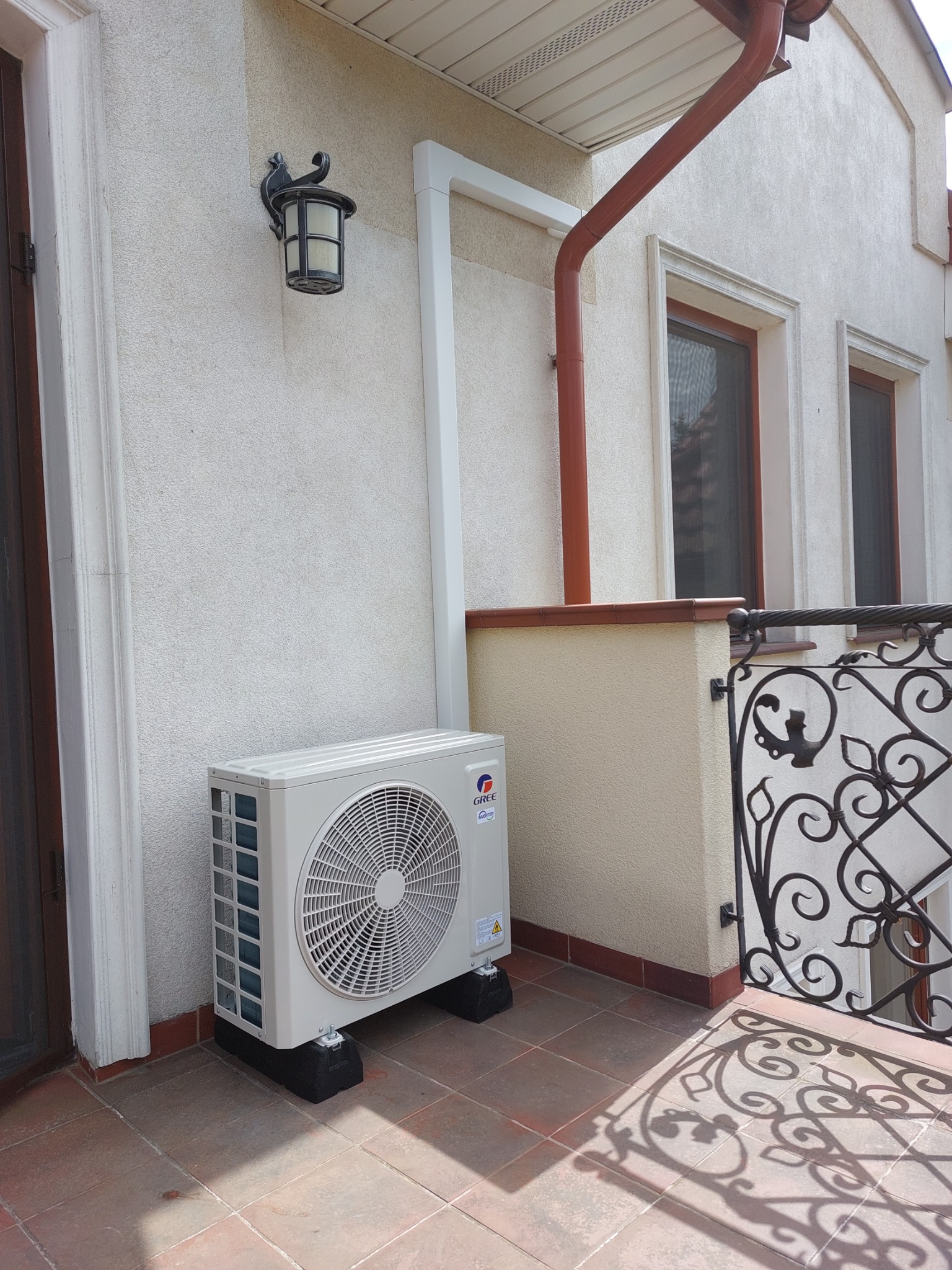 Zewnętrzna jednostka klimatyzacji marki Gree zamontowana na balkonie, z widocznymi elementami elewacji budynku, takimi jak rynna, okna i ozdobna lampa ścienna.