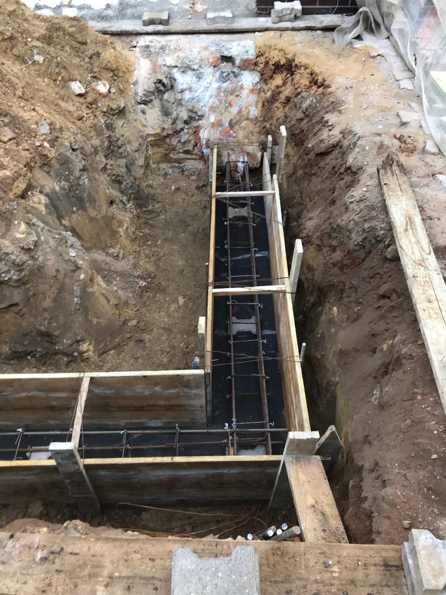 Wykop fundamentów z deskowaniem i zbrojeniem stalowym, widoczne warstwy ziemi i gruzu w wykopie.