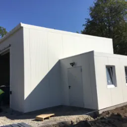 Biała hala z płyt warstwowych z drzwiami garażowymi i wejściowymi, widoczne dwie okna, pracownik w kamizelce odblaskowej wykonujący prace ziemne