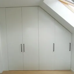 Biała szafa wnękowa z czterema drzwiami i podłużnymi, czarnymi uchwytami, dopasowana do skosu dachu, z widocznym oknem dachowym.