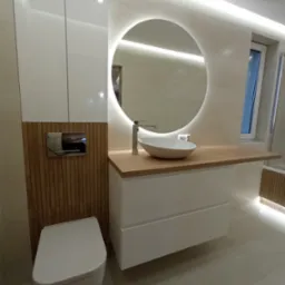 Nowoczesna łazienka z podświetlanym okrągłym lustrem, białą umywalką nablatową, drewnianym blatem, podwieszaną toaletą i szafkami w zabudowie, oświetlona listwami LED.