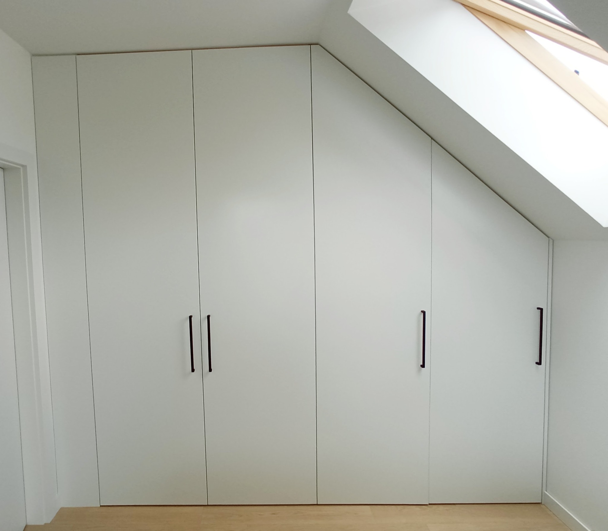 Biała szafa wnękowa z czterema drzwiami i podłużnymi, czarnymi uchwytami, dopasowana do skosu dachu, z widocznym oknem dachowym.