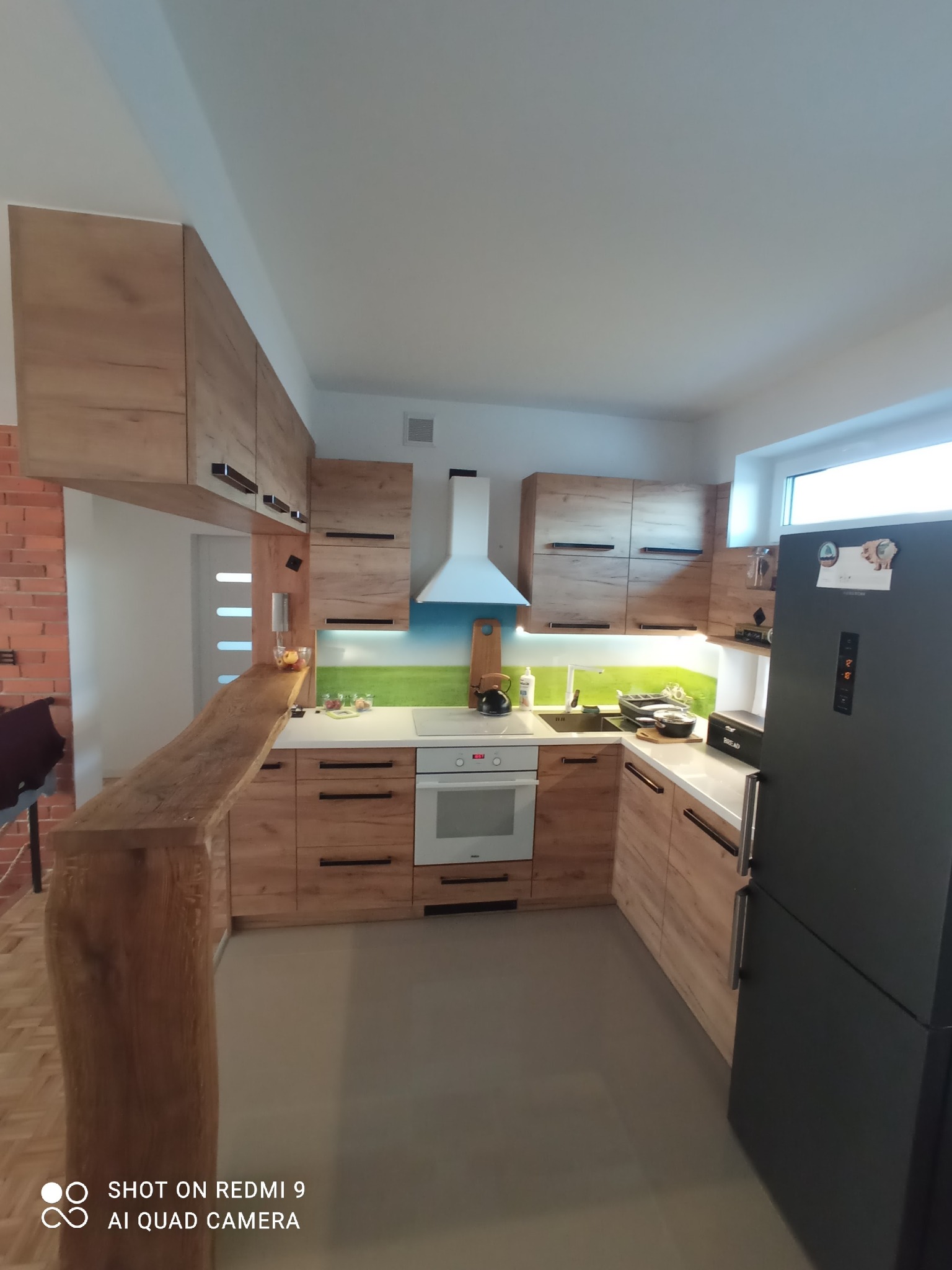 Nowoczesna kuchnia z drewnianymi frontami szafek, białym blatem i panelem szklanym z motywem łąki, widoczny również drewniany barek i czarna lodówka.