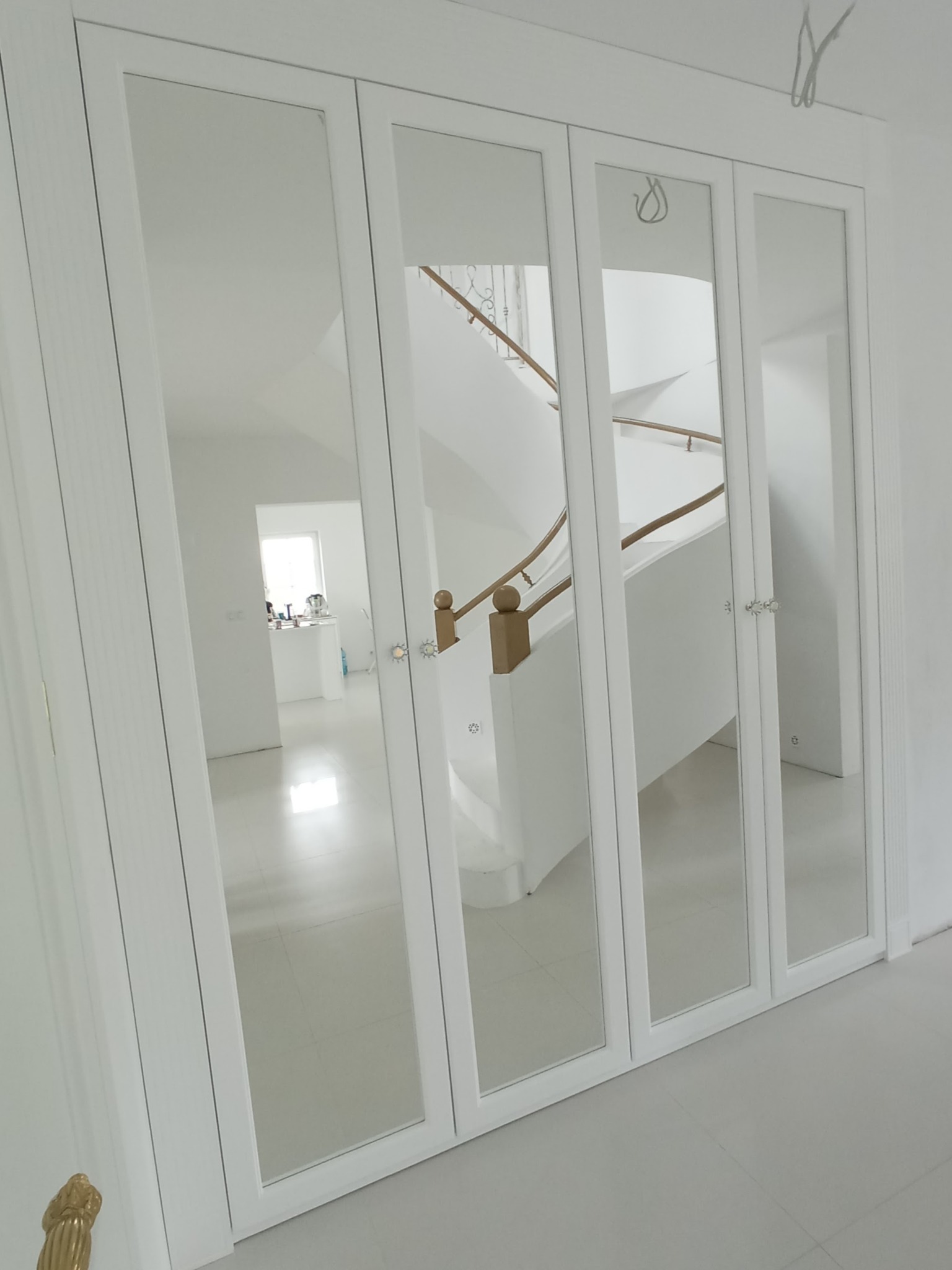 Biała szafa wnękowa z lustrzanymi drzwiami, odbijająca schody z drewnianą balustradą i jasne wnętrze domu.