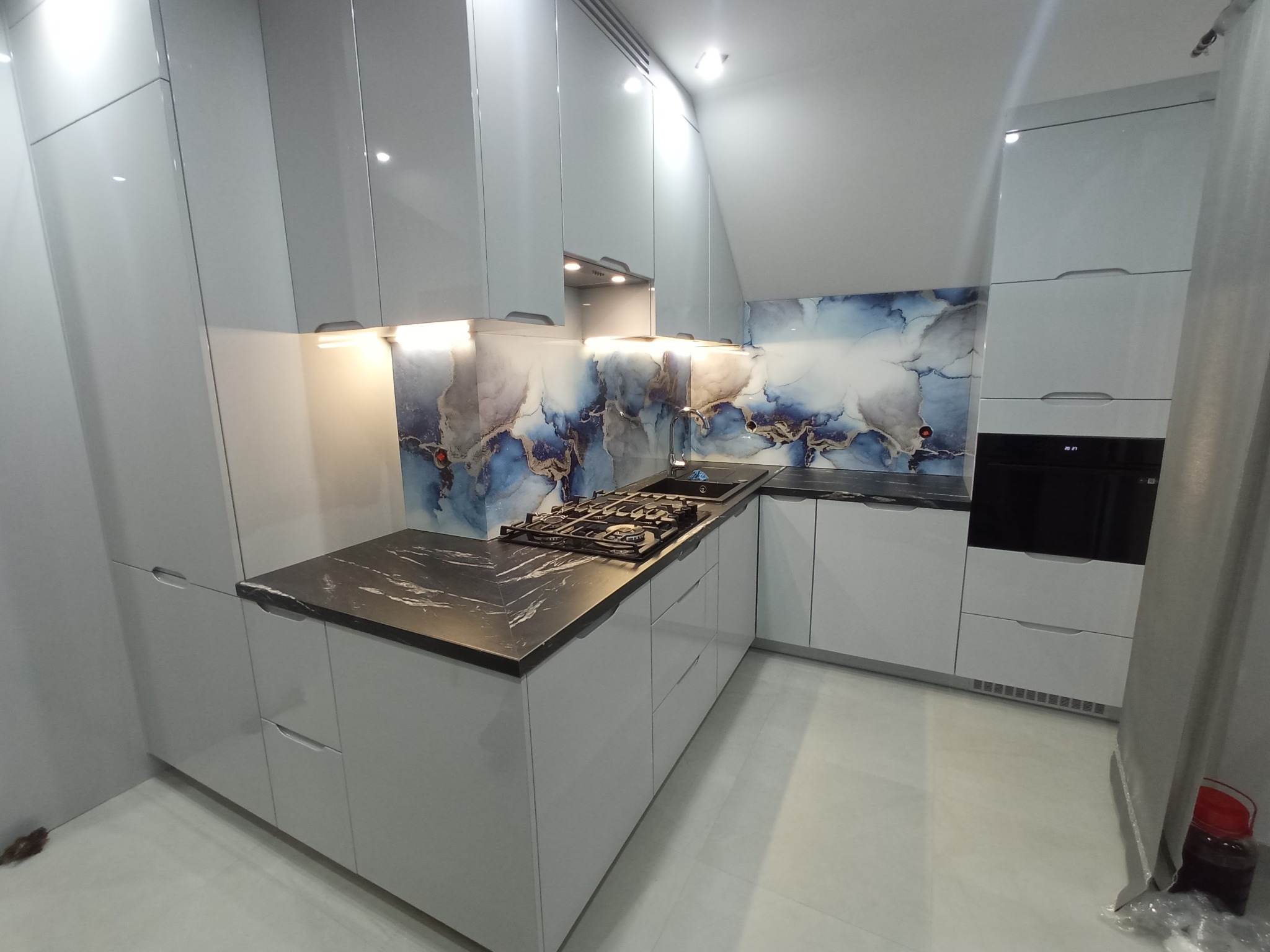 Nowoczesna kuchnia z białymi, lakierowanymi frontami, czarnym blatem z marmurkowym wzorem i dekoracyjnym panelem nad blatem w odcieniach błękitu i szarości, oświetlona listwami LED pod szafkami.