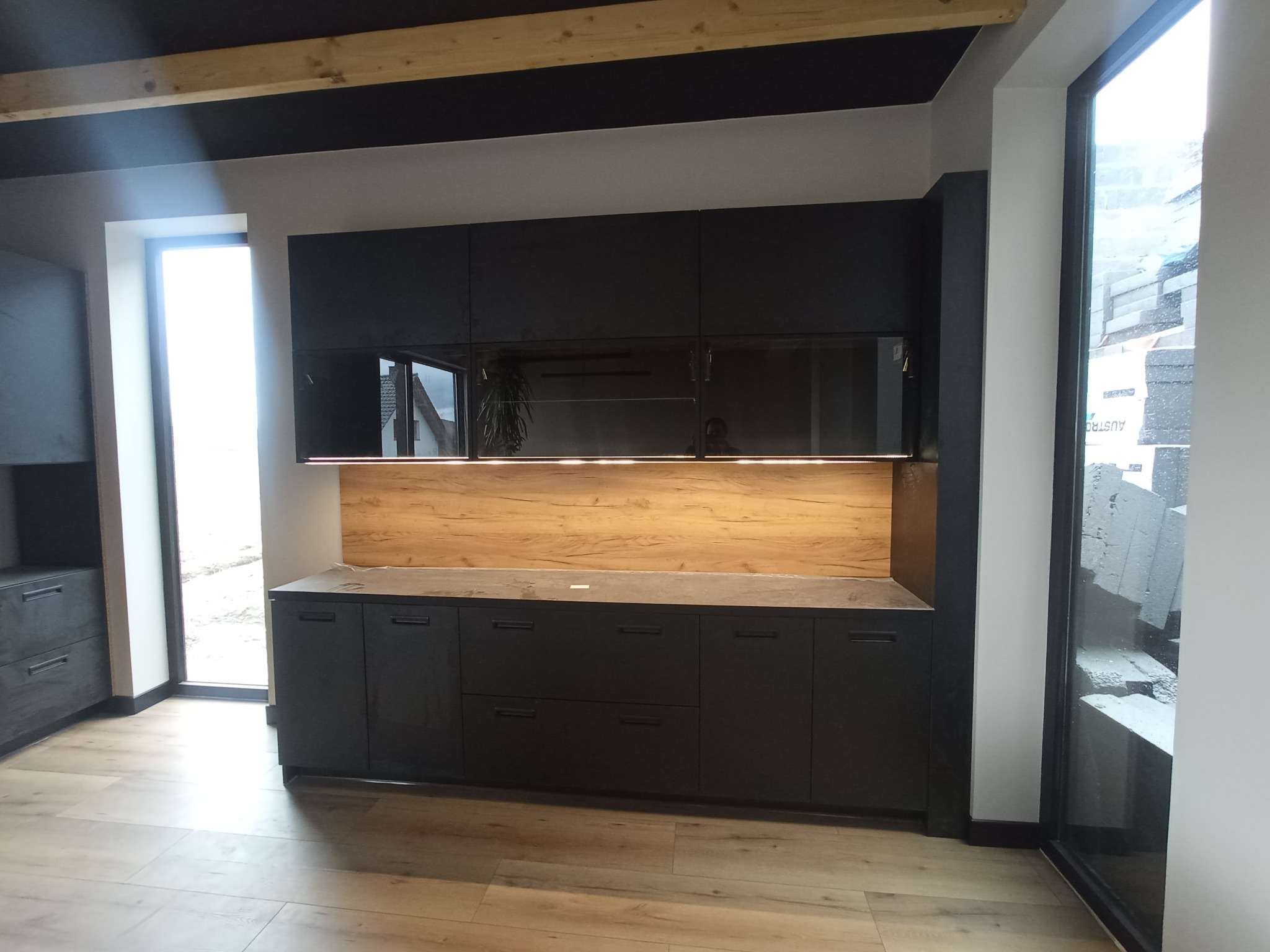 Nowoczesna, czarna zabudowa kuchenna z drewnianym panelem dekoracyjnym, oświetleniem LED i odbiciem okna w szklanych frontach szafek wiszących.