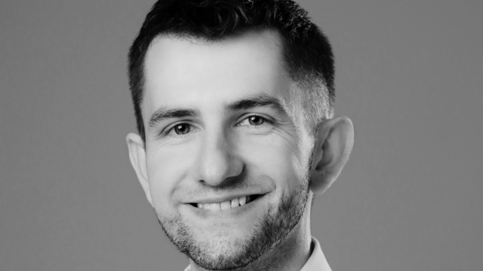 Portret uśmiechniętego mężczyzny z krótką brodą, w czerni i bieli, z wyraźnie zaznaczonymi zmarszczkami mimicznymi wokół oczu.