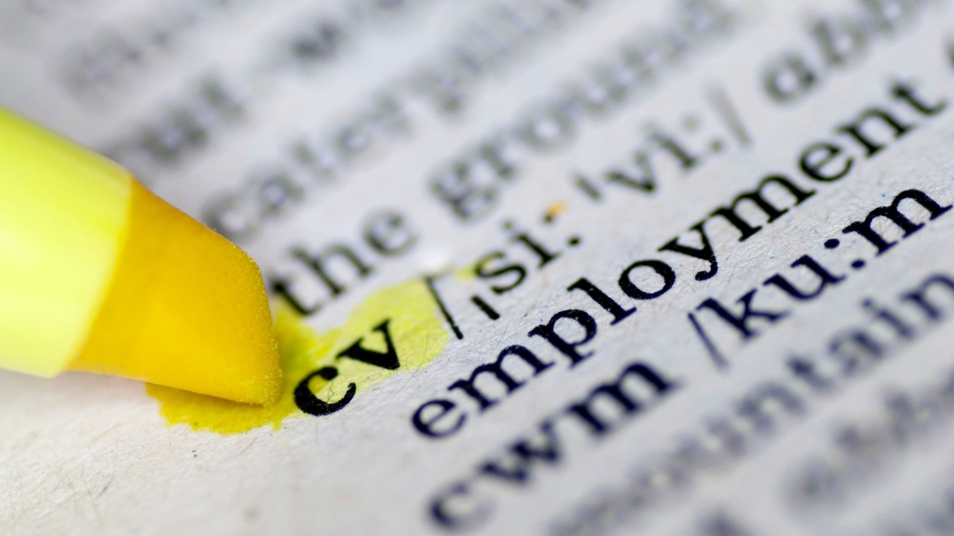 Makro ujęcie otwartej książki, gdzie tekst 'CV' jest podświetlony żółtym zakreślaczem, obok widoczny fragment słowa 'employment'.