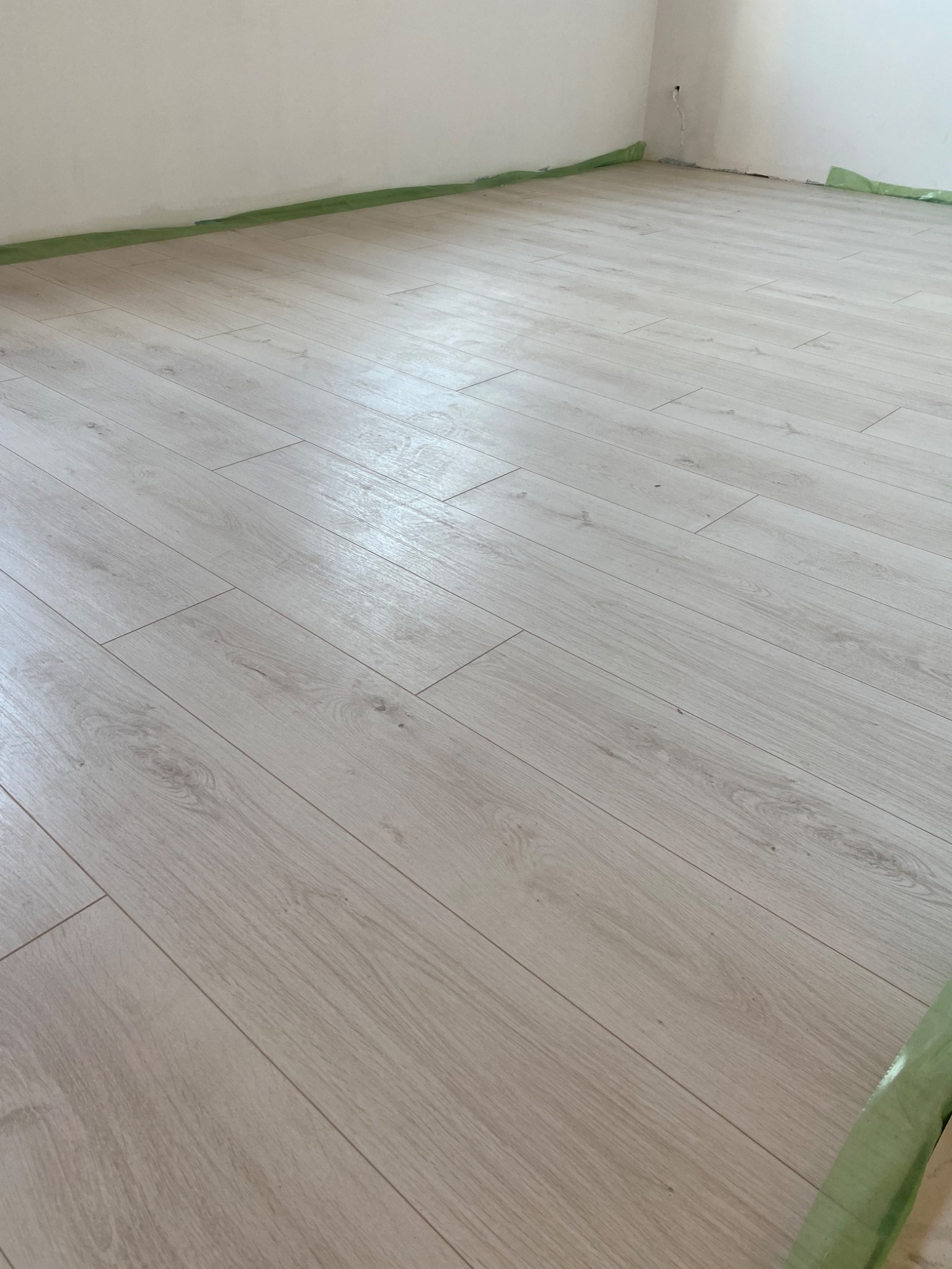 Jasne panele podłogowe ułożone w pokoju, zabezpieczone taśmą malarską przy ścianach, widoczny fragment instalacji elektrycznej.