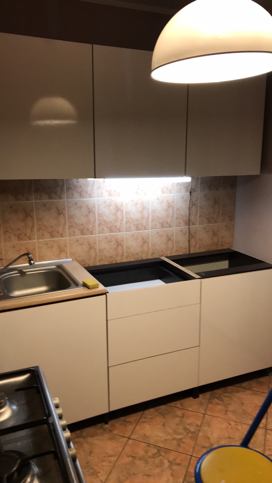Montaż białych szafek kuchennych z umywalką i miejscem na płytę grzewczą na tle beżowych płytek; widoczny fragment kuchenki i krzesła.