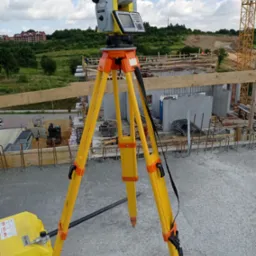 Żółty teodolit GeoMax na trójnogu, ustawiony na betonowym fundamencie budowli w trakcie wznoszenia, w tle widoczny krajobraz z budynkami i dźwigiem.