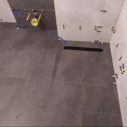 Montaż ciemnoszarych płytek podłogowych i ściennych w łazience, widoczny odpływ liniowy, żółta rura kanalizacyjna i elementy poziomujące płytki.