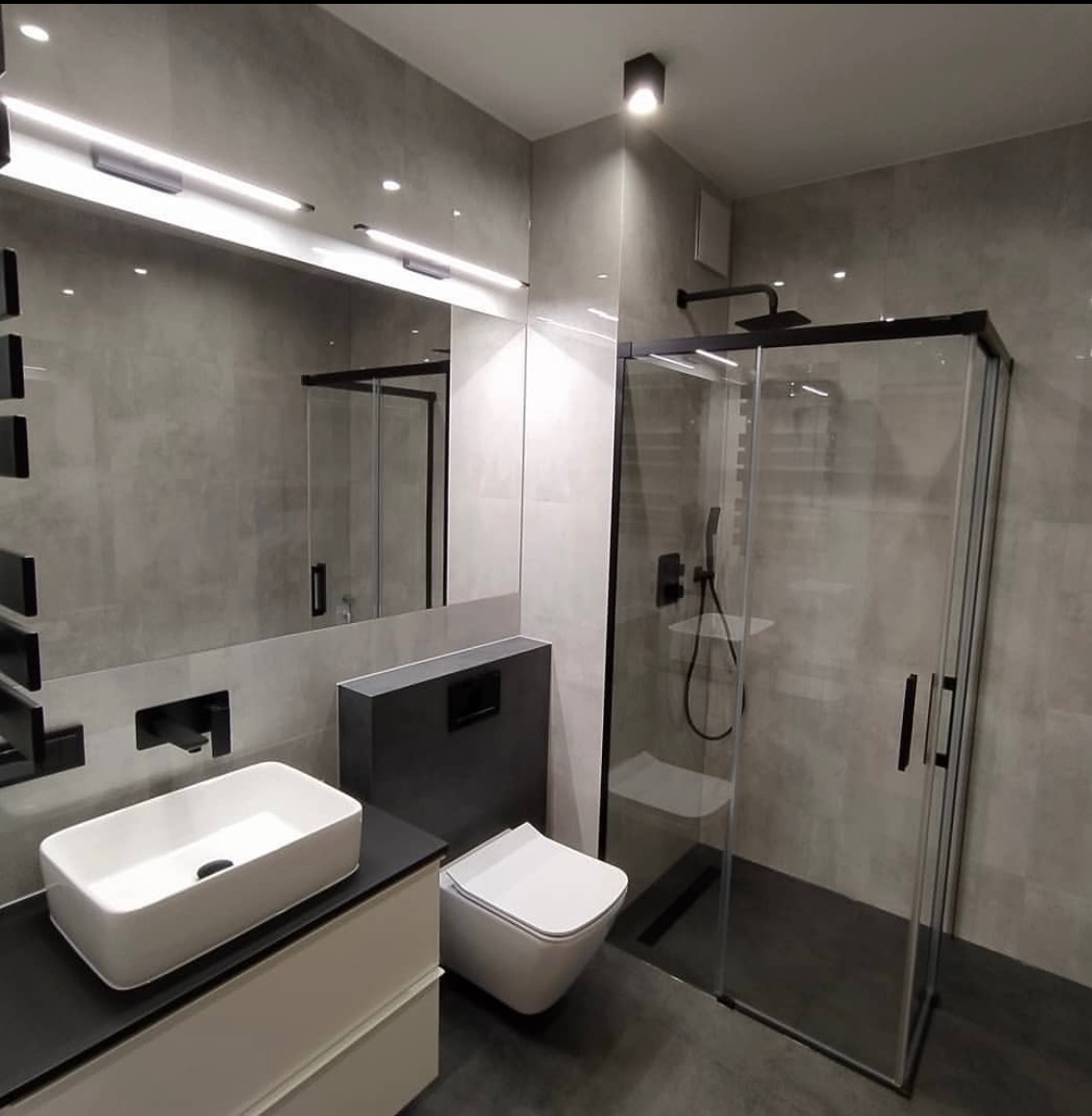 Nowoczesna łazienka w odcieniach szarości z czarnymi akcentami, kabina prysznicowa typu walk-in z czarną ramą, prostokątna umywalka nablatowa i podwieszana toaleta.