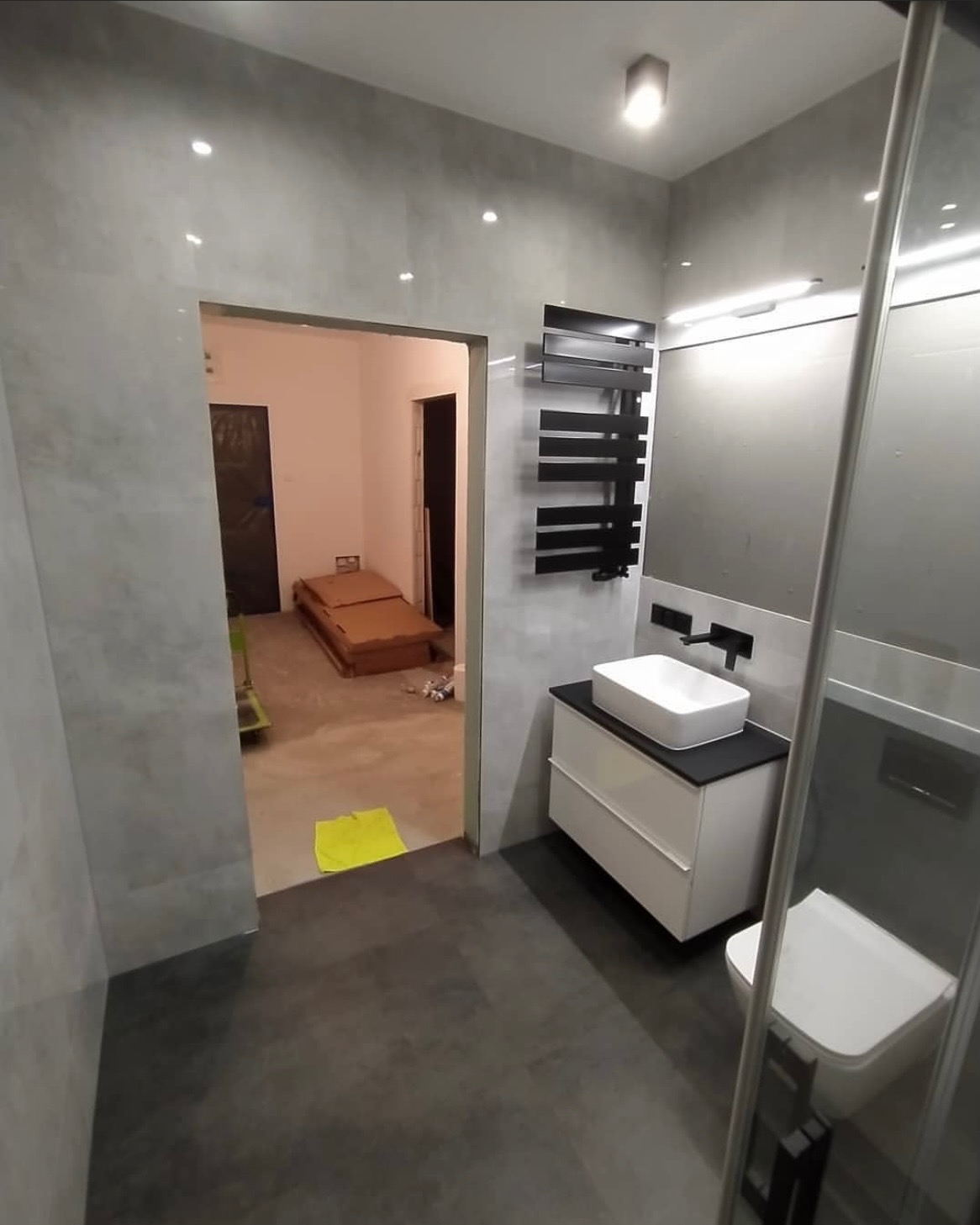 Nowoczesna łazienka w szarościach z czarnymi akcentami, widok na sąsiednie pomieszczenie z podłogą zabezpieczoną folią, żółta ścierka na progu.