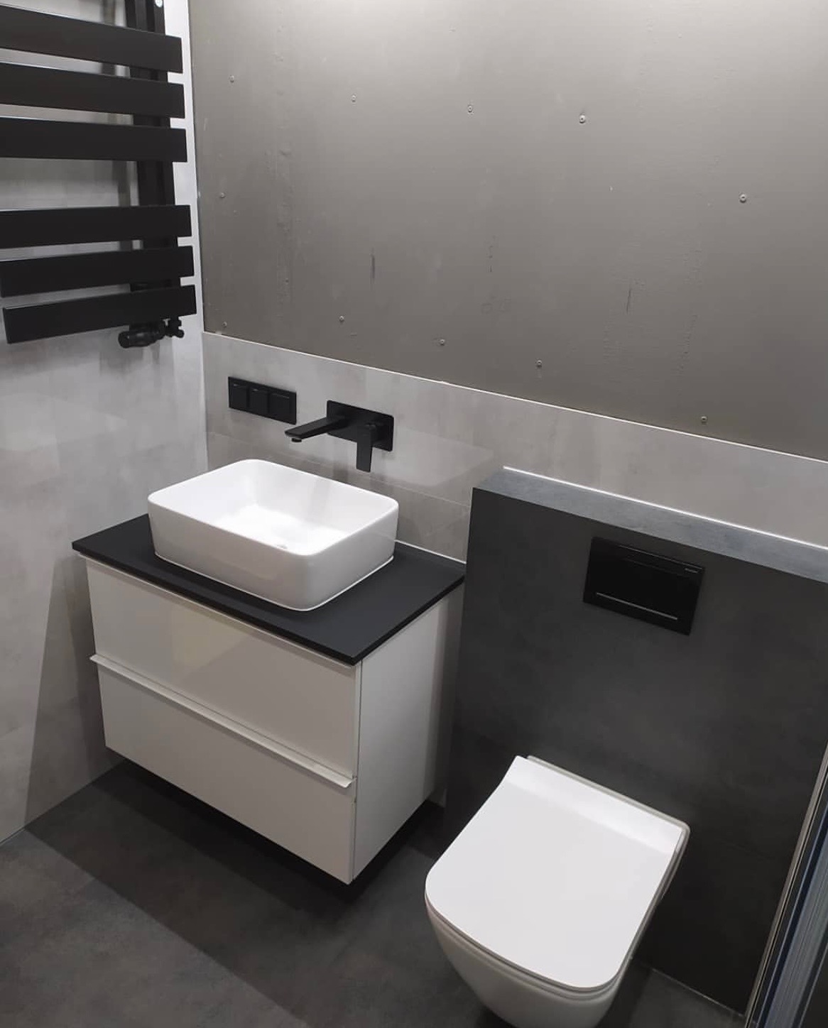 Nowoczesna łazienka w odcieniach szarości i bieli w Jaśle, z wiszącą szafką pod umywalkę z czarnym blatem, toaletą w zabudowie i grzejnikiem drabinkowym w kolorze czarnym.