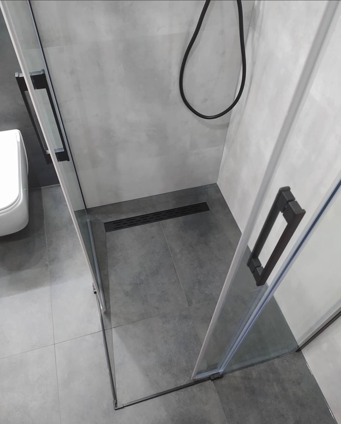 Nowoczesna kabina prysznicowa z czarnymi uchwytami i odpływem liniowym w szarej łazience, widok z góry.