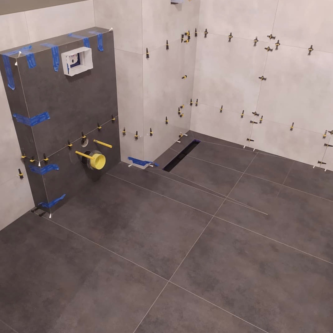 Wnętrze łazienki w trakcie wykańczania, widoczne płytki na ścianach i podłodze, system mocowania płytek, odpływ liniowy w podłodze, fragment instalacji podtynkowych zabezpieczony folią.