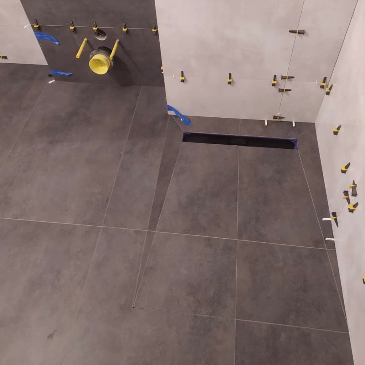 Montaż ciemnoszarych płytek podłogowych i ściennych w łazience, widoczny odpływ liniowy, żółta rura kanalizacyjna i elementy poziomujące płytki.