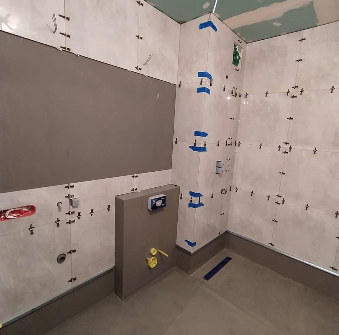 Białe płytki ścienne w trakcie układania w łazience, z użyciem krzyżyków dystansowych i niebieskiej taśmy maskującej w narożnikach, widoczna szara zabudowa stelaża WC i częściowo położona podłoga.