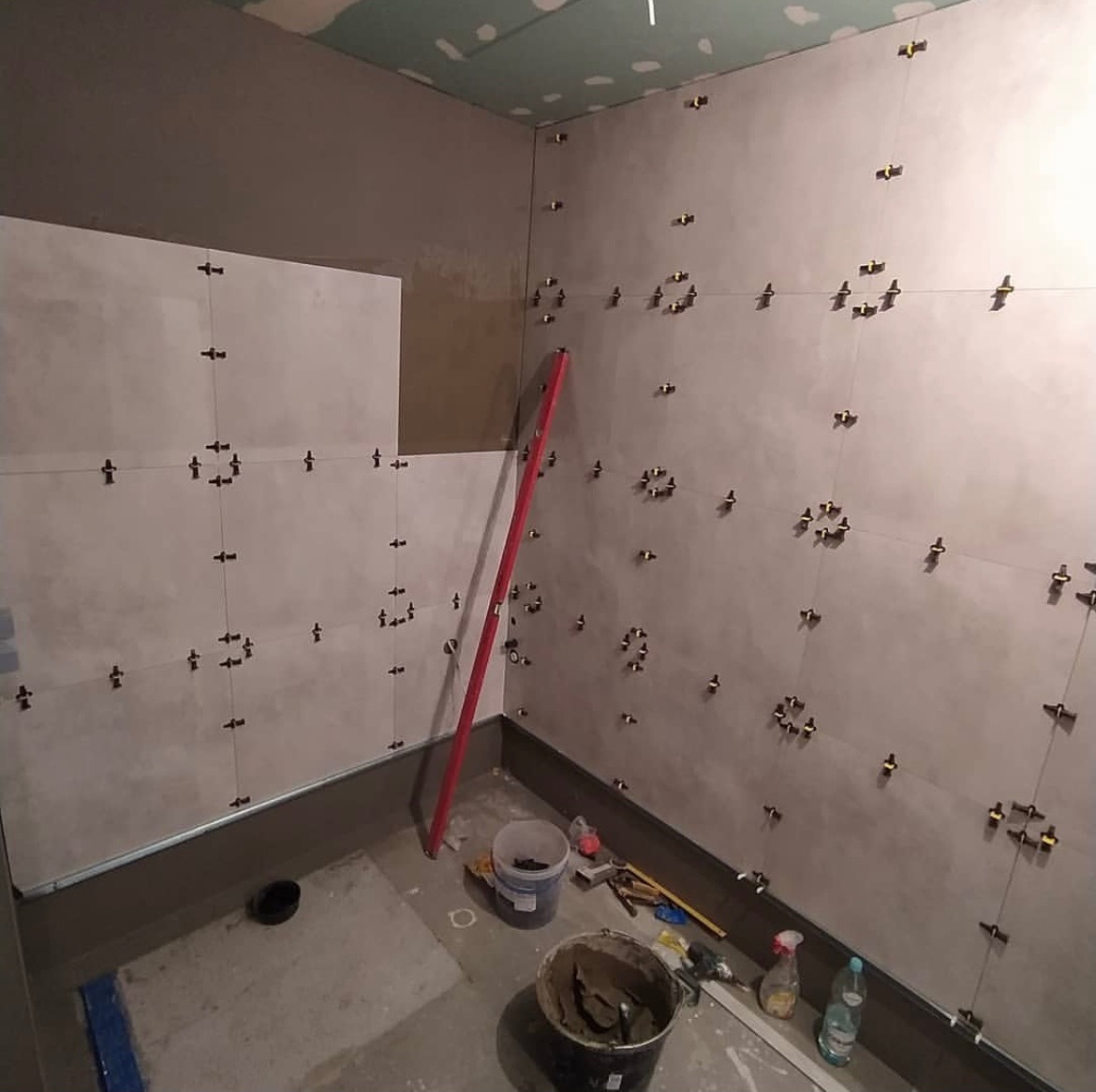 Wnętrze łazienki w trakcie wykańczania, widoczne szare płytki na ścianach, poziomica oparta o ścianę, wiadro z zaprawą i narzędzia na podłodze.