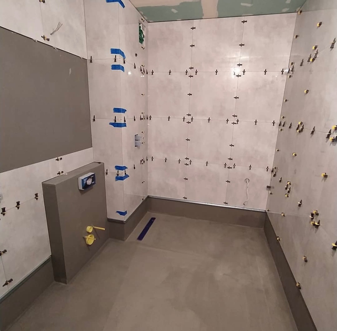 Łazienka w trakcie remontu: świeżo położone, jasne płytki z krzyżykami dystansowymi i niebieską taśmą, szara zabudowa stelaża WC, wylewka podłogowa.