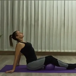 Kobieta w czarnym topie i szarych legginsach siedzi na fioletowej macie, opierając się plecami o podłogę i używając małej fioletowej piłki pod pośladkami, ćwicząc rozciąganie kręgosłupa w jasnym...