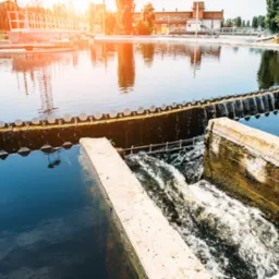 Okrągły zbiornik oczyszczalni ścieków z betonowymi elementami i przepływającą wodą, widok z góry, w tle budynek i drzewa, słoneczne odbicie na wodzie.