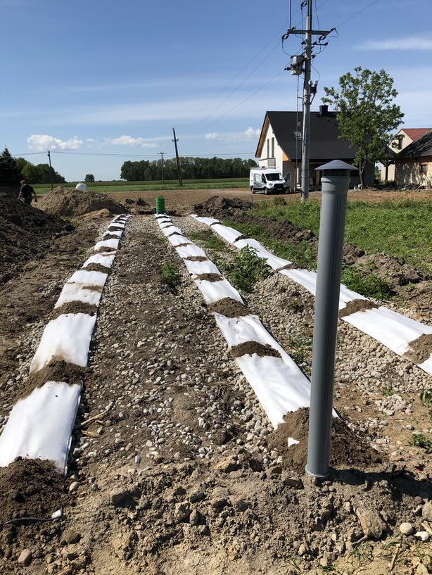 Budowa drenażu rozsączającego: rów wyłożony białą geowłókniną, obsypany żwirem i ziemią, pionowy odpowietrznik z szarego PCV, w tle dom jednorodzinny i błękitne niebo.