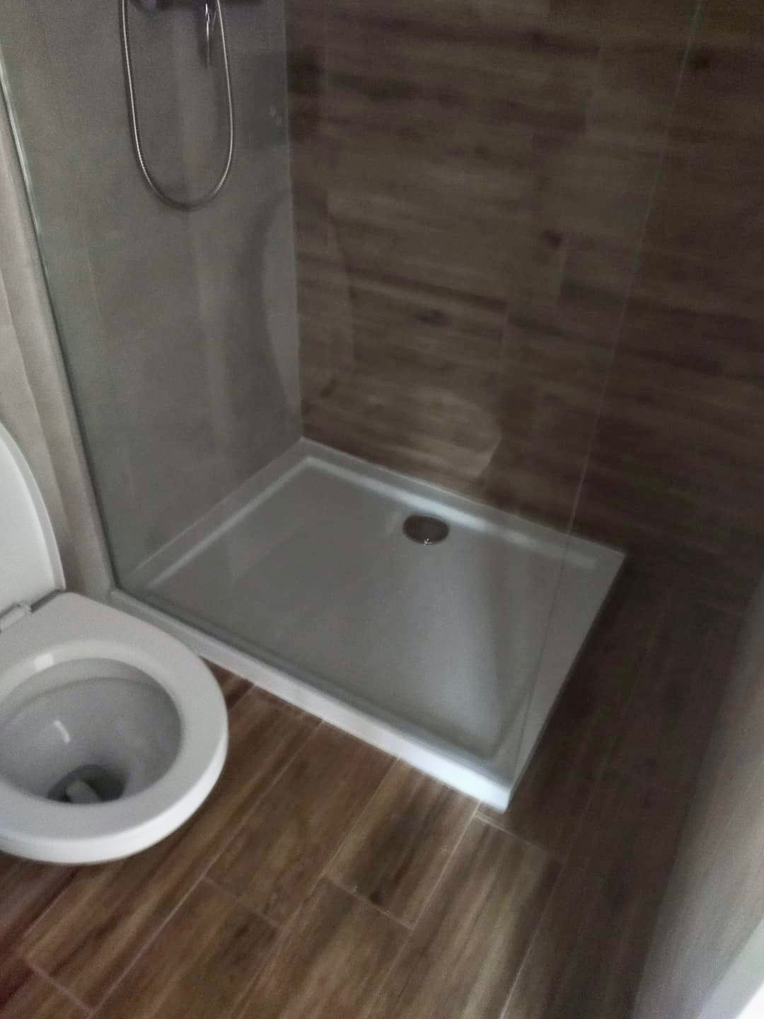 Nowoczesna kabina prysznicowa z niskim brodzikiem, wykończona płytkami imitującymi drewno, widoczny fragment toalety z otwartą klapą.