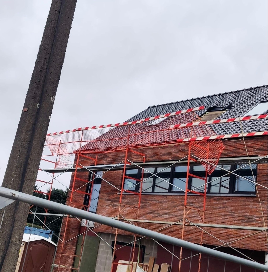 Budowa domu z czerwonej cegły na etapie montażu okien i rusztowań zabezpieczonych siatką ochronną, widoczny fragment dachu z czarnej dachówki i uszkodzeniem, słup betonowy na pierwszym planie.