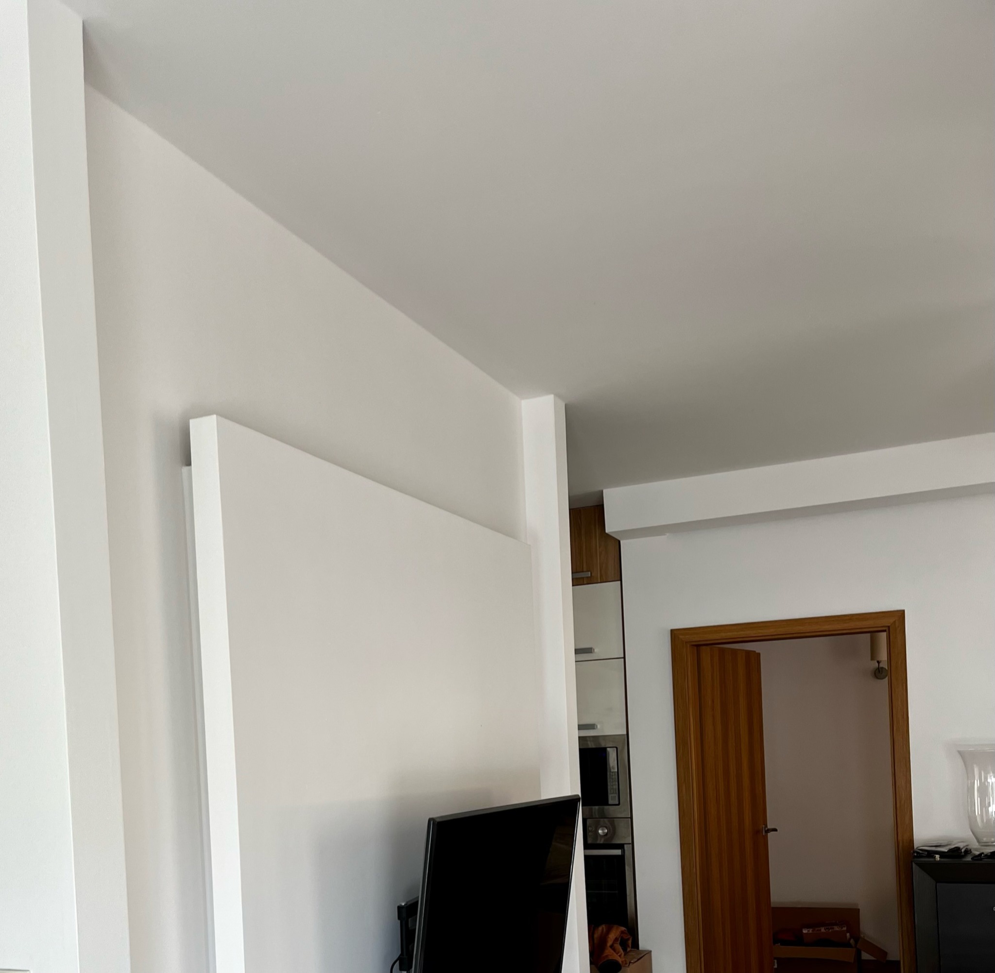 Wnętrze domu z białymi ścianami, widoczna zabudowa kuchenna w tle oraz drewniane drzwi do innego pomieszczenia.