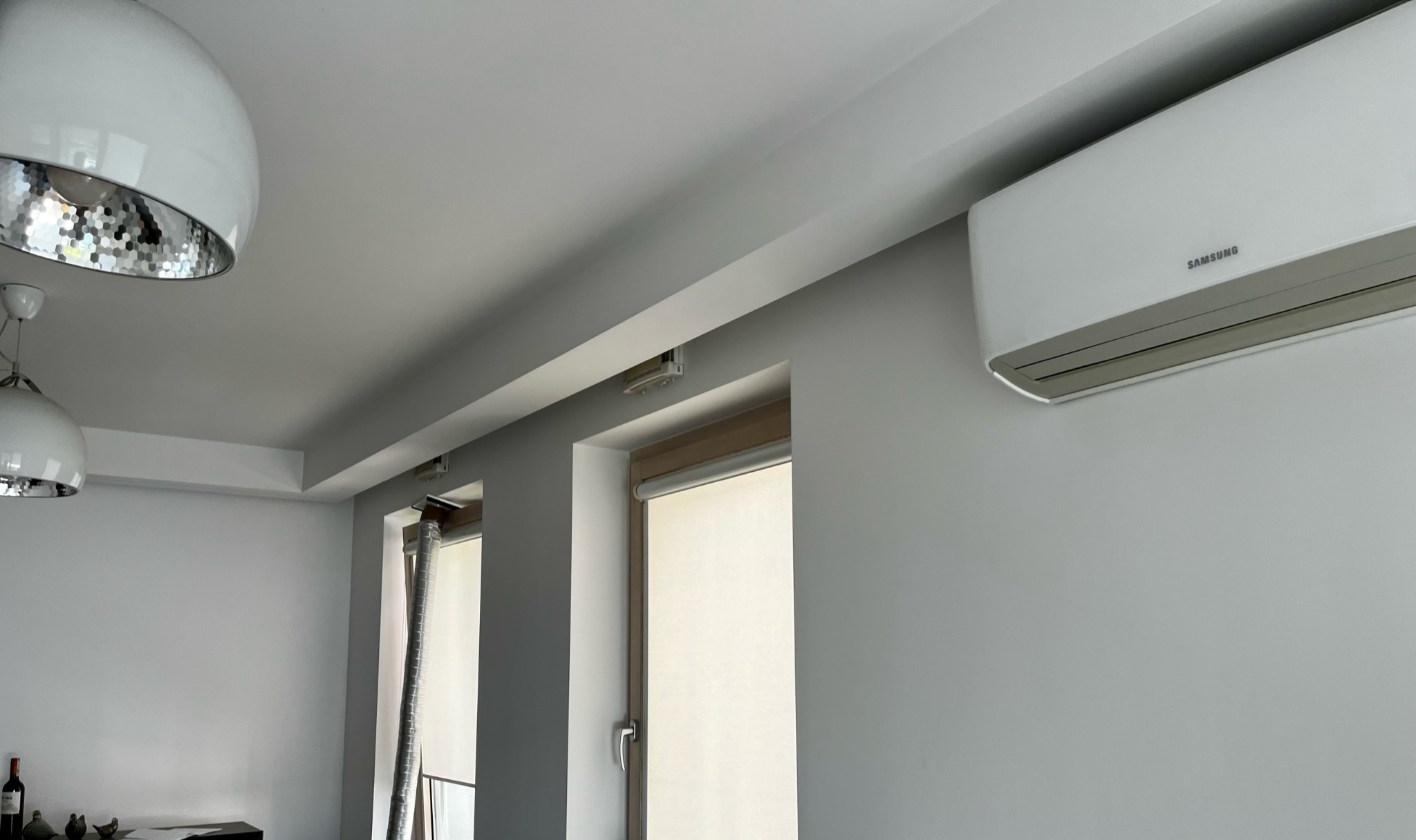 Wnętrze pokoju z klimatyzatorem Samsung na ścianie, dwoma oknami z roletami i nowoczesnymi lampami sufitowymi.