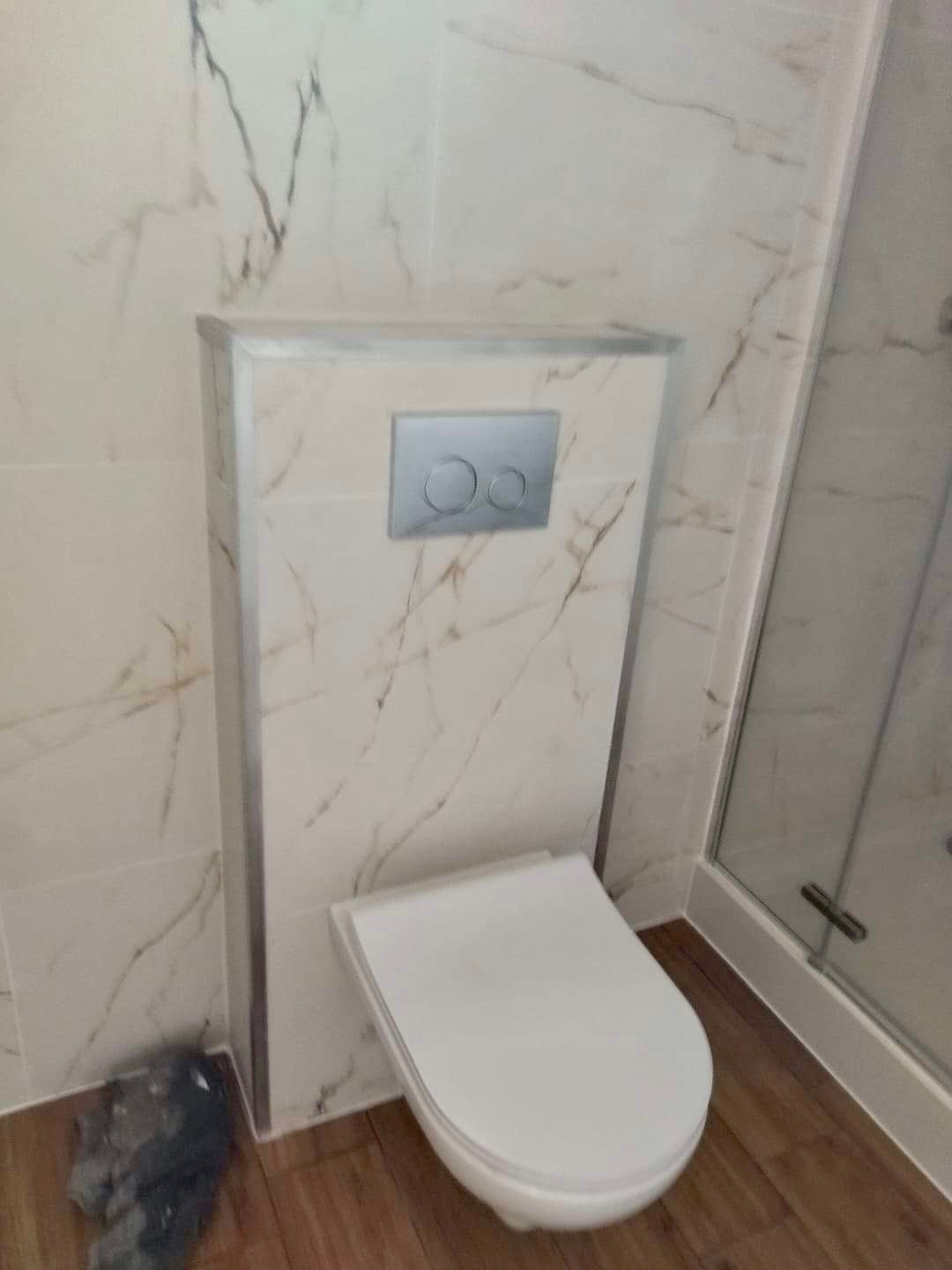 Podwieszana toaleta z marmurową zabudową i srebrnym przyciskiem spłukującym, widoczna obok kabina prysznicowa z drewnopodobną podłogą. Na podłodze widoczne zabrudzenia.