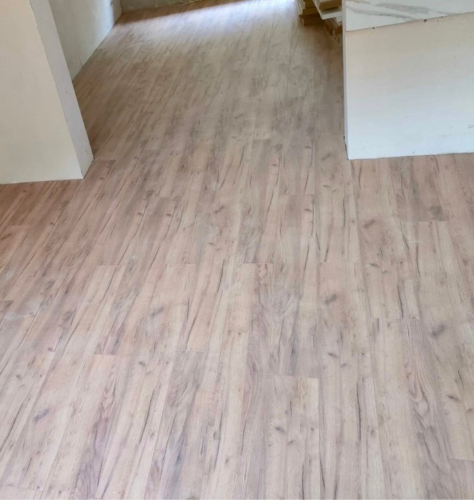 Ułożona podłoga z paneli imitujących jasne drewno, widoczne łączenia i struktura drewna, przy ścianie stos białych płyt.