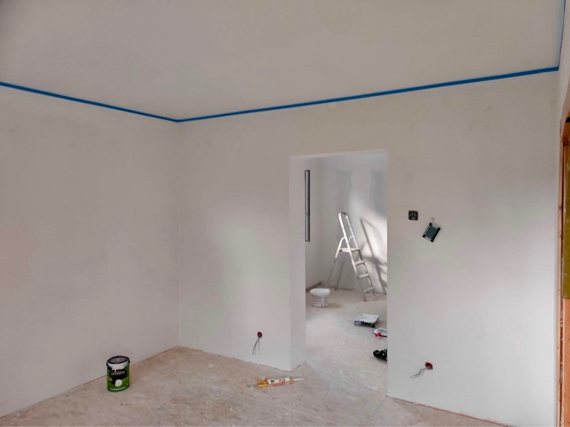 Pokój w trakcie malowania: białe ściany z niebieską taśmą malarską przy suficie, drabina, puszka farby i akcesoria malarskie na podłodze.