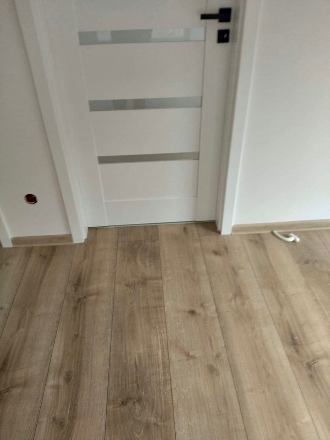 Wnętrze pokoju z panelami podłogowymi imitującymi drewno, białe drzwi z trzema poziomymi przeszkleniami i czarną klamką, widoczny fragment ściany z gniazdkiem elektrycznym.