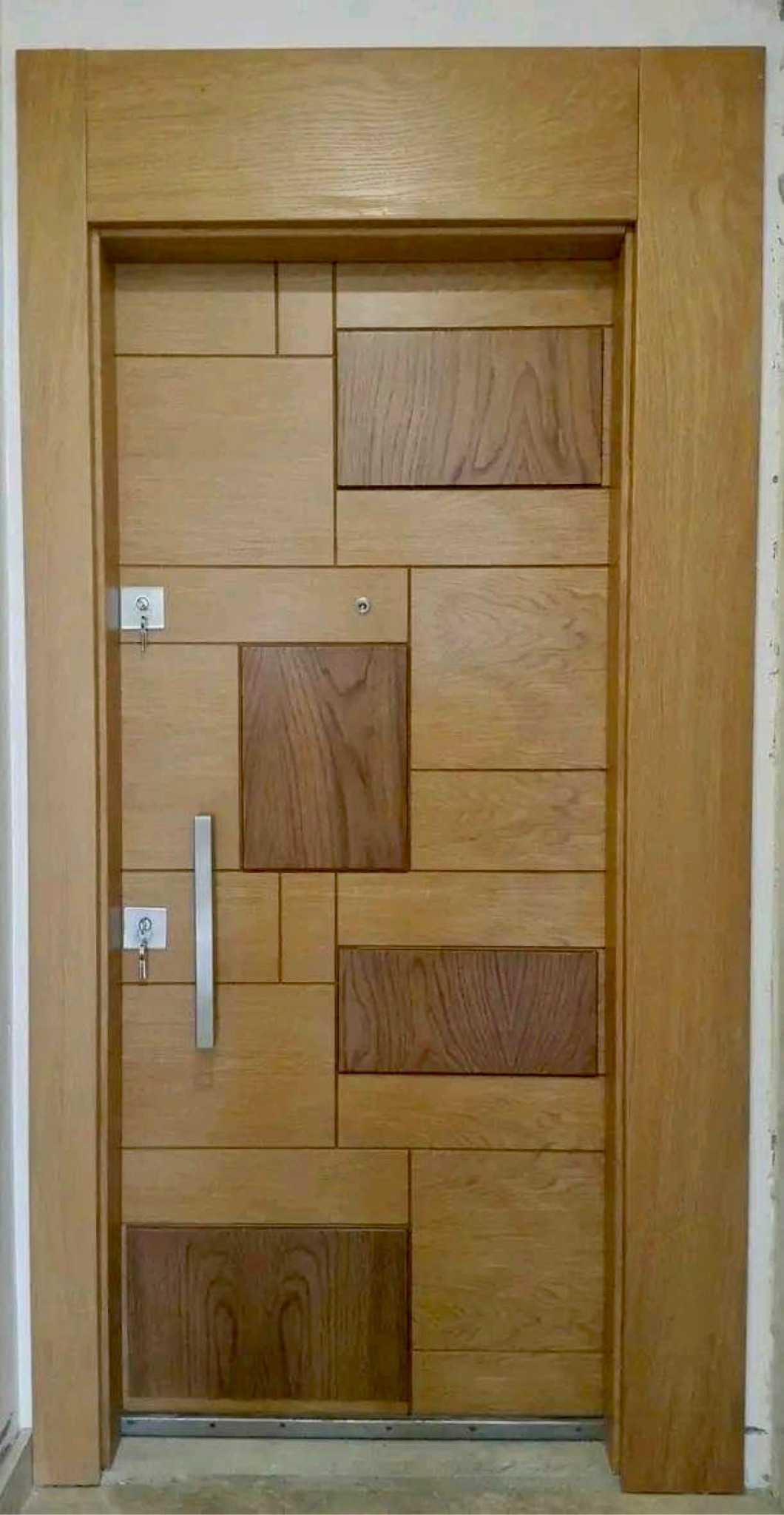 Drewniane drzwi wejściowe z geometrycznym wzorem z różnych odcieni drewna, wyposażone w srebrną klamkę i dwa zamki z kluczami.