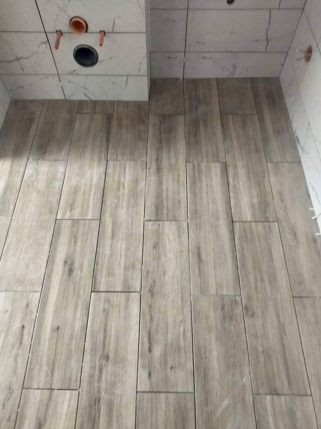 Ułożone podłużne płytki podłogowe imitujące drewno w łazience, widoczne podejścia instalacyjne do armatury na ścianie wyłożonej białymi płytkami imitującymi marmur.