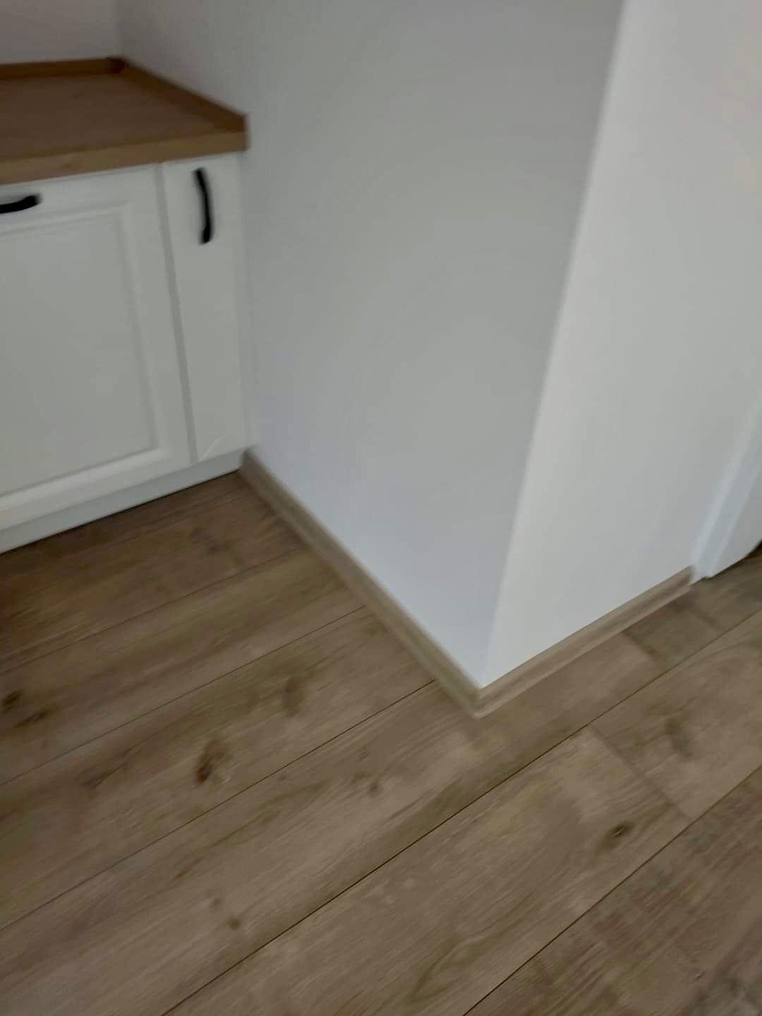 Fragment wnętrza: laminowana podłoga w odcieniach brązu, biała listwa przypodłogowa i biała szafka z blatem w kolorze drewna w rogu pomieszczenia.