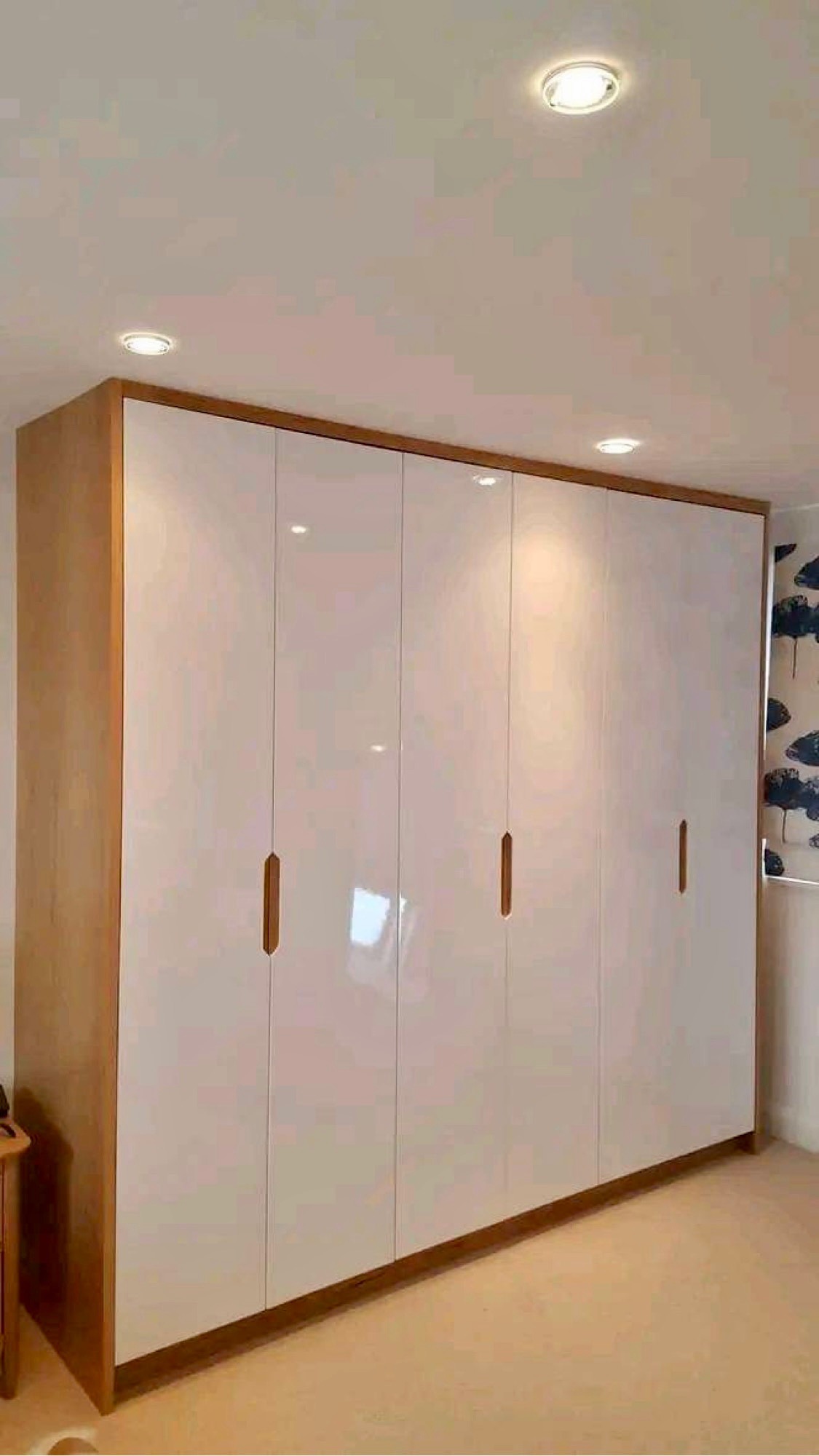 Duża szafa wnękowa z białymi, lakierowanymi frontami i drewnianym obramowaniem, oświetlona trzema punktowymi lampami sufitowymi.