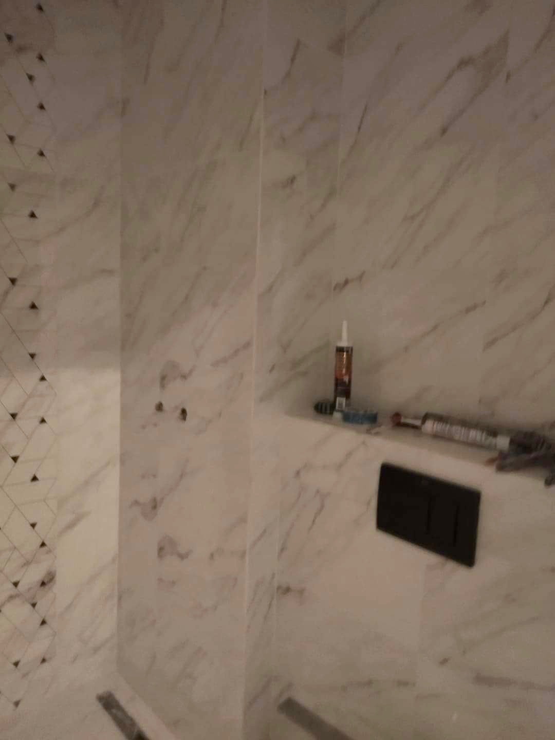 Wykończona łazienka z marmurowymi płytkami, widoczna czarna armatura, silikon i narzędzia na półce w ścianie.