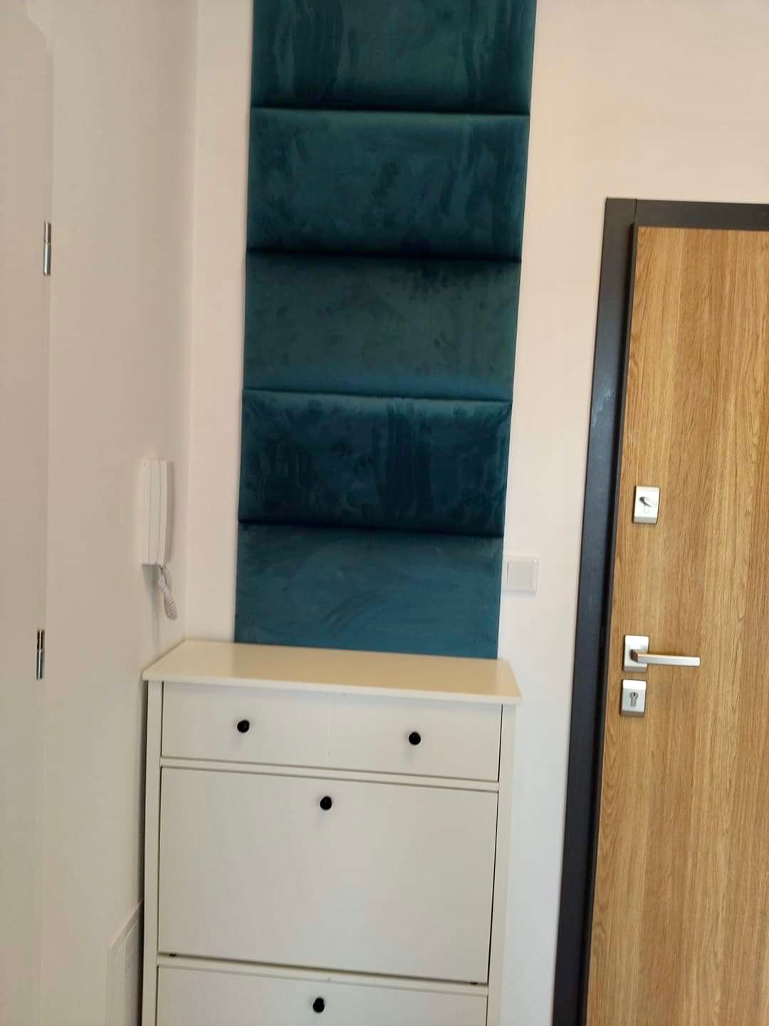 Panel ścienny z zielonego weluru, zamontowany pionowo obok białej komody na buty i drzwi z drewnianą okleiną w nowoczesnym wnętrzu.